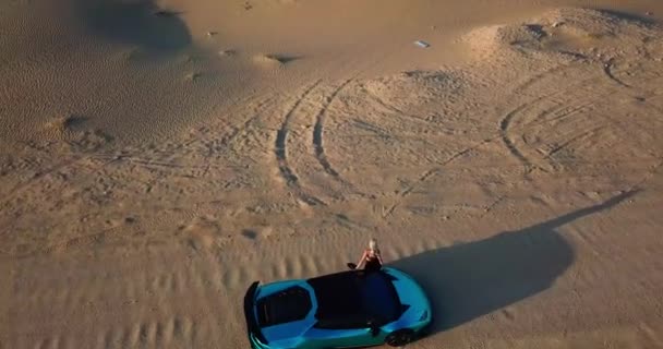 无人机抓住了一个穿着轻便衣服的年轻女子 她站在一辆昂贵的跑车旁边 坐在沙漠的沙滩上 空中景观 慢动作 — 图库视频影像