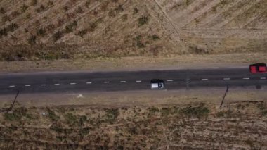 İnsansız hava aracı bozkır ve çalılarla çevrili bir kır yolunun üzerinde uçuyor. Yol boyunca farklı renklerde arabalar sürüyor. Hava görünümü