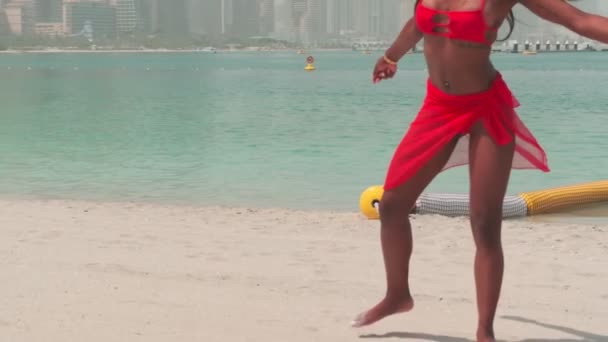 ビキニを着た若い女と赤いドレスは 波の水の中の砂浜を裸足で歩く スローモーション — ストック動画