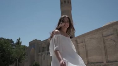 Açık beyaz elbiseli, uzun dalgalı saçlı genç bir kadın antik bir minarenin önünden geçiyor. Yavaş Hareket.
