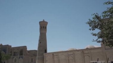 Genç bir kadın açık beyaz bir elbiseyle dalgalı saçlarıyla antik bir minarenin yanındaki açık bir göletin önünden geçiyor. Yavaş Hareket, Özbekistan.