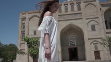Açık beyaz elbiseli ve sandaletli, dalgalı saçlı genç bir kadın antik bir doğu şehrinde yürüyor. Yavaş Hareket, Özbekistan