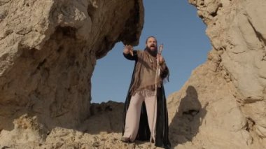 Genç sakallı bir şaman çölün kum tepelerinin arasında bir kaya mağarasında dururken dua eder. Yavaş çekim
