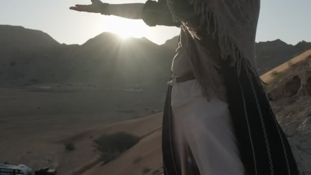 砂漠の砂丘の間に沈む夕日を背景に ひげを生やした若いシャーマンが祈ります スローモーション アラブ首長国連邦 — ストック動画
