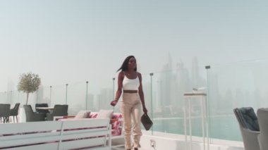 Dubai, BAE 'ye tepeden bakan cam bir balkonda beyaz takım elbiseli esmer bir kadın. Yavaş Hareket.