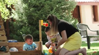 Annesi ve kardeşiyle birlikte küçük bir çocuk çimenlikteki ahşap bir kuşhanede renkli toplarla oynuyorlar. Yaz, güneşli bir gün. Yavaş çekim, Yakın çekim.