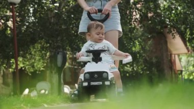 Şortlu genç bir anne, küçük oğlunu oyuncak bir arabaya bindiriyor. Park, güneşli bir gün. Yavaş Hareket.