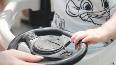 Şortlu genç bir anne, küçük oğlunu oyuncak bir arabaya bindiriyor. Park, güneşli bir gün. Yavaş Hareket, Kapat.