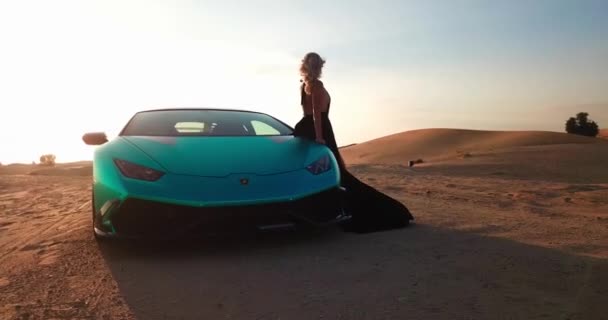 ドローンは 砂漠の砂の中に高価なスポーツカーの隣に立っている軽いドレスで若い女性をキャプチャします エアリアルビュー スローモーション — ストック動画