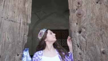 Uzun saçlı, kısa beyaz elbiseli ve ahşap bir kapının arkasında oryantal cüppeli genç bir kadın. Yavaş Hareket, Özbekistan