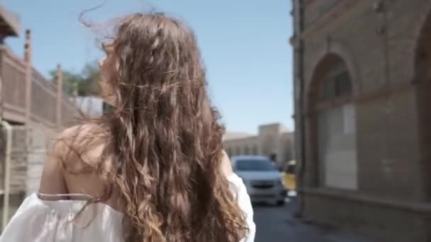 一位身穿浅白太阳衣和凉鞋 头发飘扬的年轻女子穿过古老的东部城市 乌兹别克斯坦慢动作 — 图库视频影像