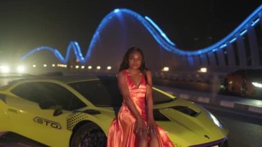 Gece vakti aydınlık bir köprüde, spor bir arabanın kurumu üzerinde oturan, gece kıyafetli esmer bir genç kadın. Slow Motion, BAE, Dubai.