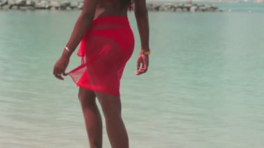 Bikinili esmer ve açık kırmızı elbiseli genç bir kadın sörf suyunda kumlu bir sahilde yalınayak yürüyor. Yavaş çekim