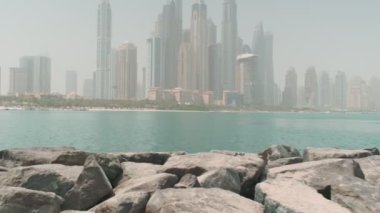 Bikinili esmer ve açık kırmızı elbiseli genç bir kadın denizin ve Dubai 'nin gökdelenlerinin önünde çıplak ayakla kayalarda yürüyor. BAE, Ağır çekim.