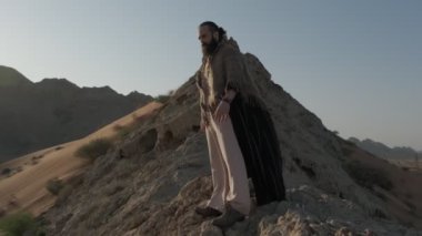 Genç sakallı bir şaman çölün kum tepeleri arasında gün batımının arka planına karşı dua ediyor. Ağır çekim, BAE