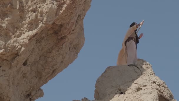 白い服を着た若い髭を生やしたシャーマンが 澄んだ空を背景に岩の上に立って祈る スローモーション アラブ首長国連邦 — ストック動画