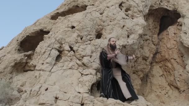 一个拿着手杖和萨满衣服的人站在岩石上 站在沙漠的沙滩上跳舞 晴朗的天空 缓慢的运动 — 图库视频影像