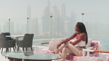 Dubai, BAE 'ye tepeden bakan cam bir balkonda beyaz takım elbiseli esmer bir kadın. Yavaş Hareket.