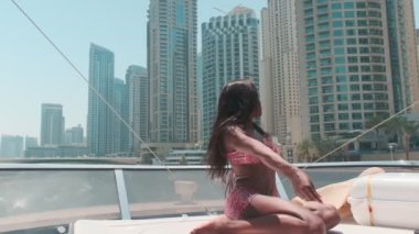 Dubai gökdelenlerinin arka planına karşı özel bir teknenin güvertesinde kısa parlak elbiseli esmer bir kadın. BAE, Ağır çekim
