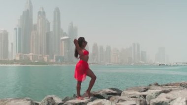 Bikinili esmer ve açık kırmızı elbiseli genç bir kadın denizin ve Dubai 'nin gökdelenlerinin önünde çıplak ayakla kayalarda yürüyor. BAE, Ağır çekim.