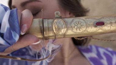 Uzun dalgalı saçlı, doğu cübbesi ve şapkası olan genç bir kadın, antik bir şehrin arka planına karşı Asya kılıcını tutuyor.