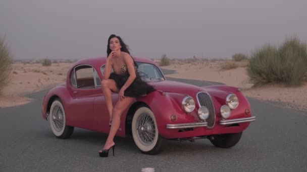 黒いドレスとハイヒールの靴の若い壮観な女性は 砂漠の真ん中に赤いヴィンテージカーの隣に道路に立っています スローモーション — ストック動画