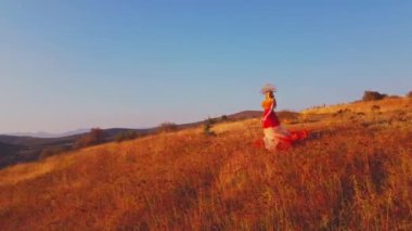 Drone, sonbahar kıyafetiyle günbatımı arka planında dağlardaki çayırlarda genç bir kadının üzerinden uçuyor. Hava görünümü.