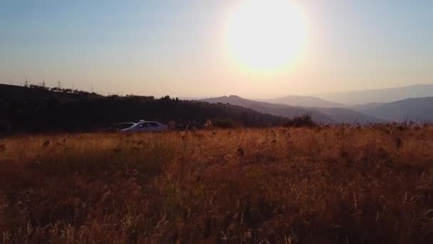 ドローンは 日没の背景にある山の草地に秋の服を着たフィルムクルーと若い女性の上を飛ぶ エアリアルビュー — ストック動画