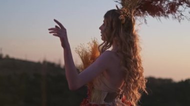 Dikenli başlıklı ve sonbahar çiçekli elbisesi içinde genç bir kadın, elinde yapraklarla elinde kuru dallarla dağlardaki günbatımının arka planında duruyor. Yavaş Hareket