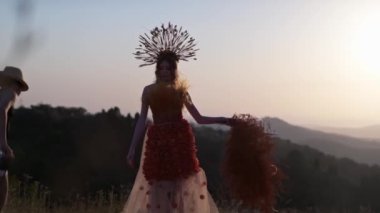 Film ekibi, başlıklı, mısır kulaklı, sonbahar elbisesiyle, çiçeklerden ve yapraklardan yapılmış genç bir manken hazırlıyor dağlarda gün batımında. Yavaş Hareket.