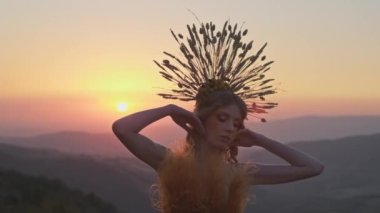 Dikenli başlıklı genç bayan manken ve dağlarda günbatımının arka planında çiçeklerden ve otlardan yapılmış elbise. Yavaş Hareket.