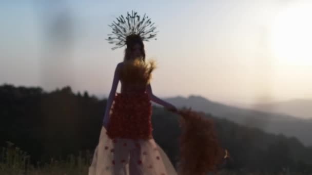 花とトウモロコシの耳で作られたヘッドドレスで作られたデザイナーのドレスの若い女性モデルは 山の夕日を背景に立っています 秋の花を持っているし — ストック動画