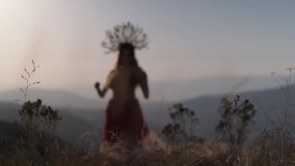 デザイナーのヘッドドレスの若い女性モデルは 山の夕日の背景に花と草で作られたドレスで作られています スローモーション — ストック動画