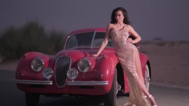 Uzun saçlı ve gece elbisesi giyen genç bir kadın pahalı bir klasik kırmızı arabanın yanında yüksek topuklu ayakkabılar içinde duruyor. Yavaş Hareket.