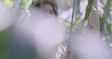 A young beautiful girl in a wreath of flowers swings on a swing among a green garden. Slow motion