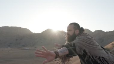 Elinde asası ve şaman kıyafetleri olan bir adam çölün kumları arasında bir kayanın üzerinde duruyor ve dans ediyor. Gökyüzü açık, yavaş çekim