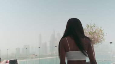 Dubai, BAE 'ye tepeden bakan cam bir balkonda beyaz takım elbiseli esmer bir kadın. Yavaş Hareket.