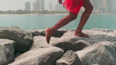 Bikinili esmer ve açık kırmızı elbiseli genç bir kadın denizin ve Dubai 'nin gökdelenlerinin önünde çıplak ayakla kayalarda yürüyor. BAE, Ağır çekim.