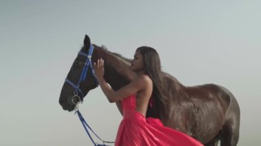 Kırmızı elbiseli genç esmer bir kadın okyanus kıyısında bir atın yanında duruyor. Yavaş çekim.
