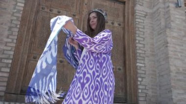 Uzun saçlı, kısa beyaz elbiseli ve ahşap bir kapının arkasında oryantal cüppeli genç bir kadın. Yavaş Hareket, Özbekistan