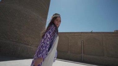 Beyaz elbiseli ve uzun doğulu cüppeli genç bir kadın eski bir minarenin yanında gün ışığında dans ediyor. Yavaş Hareket, Özbekistan