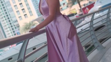 Dalgalı beyaz saçlı ve uzun leylak elbiseli genç bir kadın Dubai 'deki yatların ve gökdelenlerin önünde rıhtımda duruyor. Yavaş Hareket