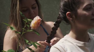 Genç bir kadın, kız kardeşlerinin uzun saçlarını evin avlusunda örüyor. Güneşli bir gün, çiçek açan bitkiler. Yavaş Hareket.