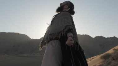 Genç sakallı bir şaman çölün kum tepeleri arasında gün batımının arka planına karşı dua ediyor. Ağır çekim, BAE