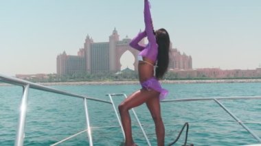 Beyaz bikinili esmer bir kadın ve şehrin arka planına karşı özel bir teknede açık renk bir elbise giyiyor. Ağır çekim, BAE