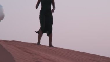 Uzun siyah elbiseli esmer bir genç kadın ve bir Arap atkısı çöl kumunda çıplak ayakla kumsalda duran beyaz bir SUV 'ye doğru yürüyor. Yavaş çekim