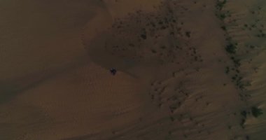 Bir dron, Arap giysileri içinde çölün kum tepesinde oturan bir adamın üzerinden gökyüzünün arka planına doğru uçuyor. Hava Görünümü.