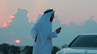 Ulusal Arap kıyafetleri içinde sakallı genç bir işadamı pahalı bir arabanın yanında telefonla konuşuyor. Arka planda gün batımı bulutlu gökyüzü.