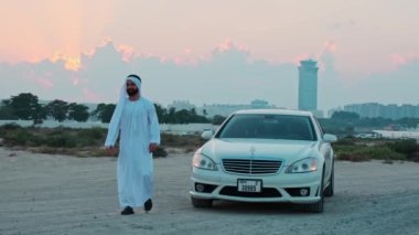 Ulusal Arap kıyafetleri içinde sakallı genç bir işadamı gün batımında pahalı bir lüks arabanın yanında dans ediyor. Dubai gökdelenlerinin arka planında