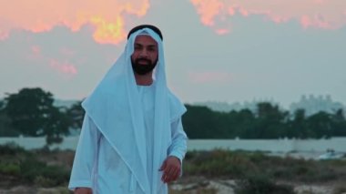 Ulusal Arap kıyafetleri içinde sakallı genç bir işadamı gün batımının arka planında dans ediyor. Dubais gökdelenleri ufukta görünüyor..